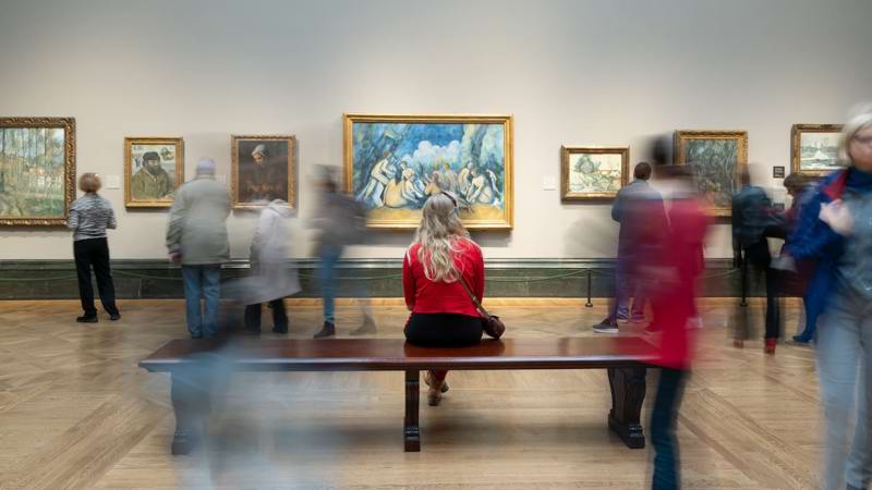 Timelapse Blur Of Woman Sitting On Bench Listening To Audioguide Looking At Painting